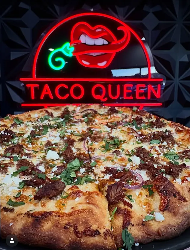 Birria pizza topped with shredded beef, red onion, cilantro and cotija under the Taco Queen neon sign